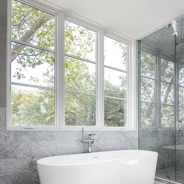 Bright modern bathroom featuring a white soaking tub set against gray marble tile beneath a large, three-panel white casement window with glass grids looking out at green trees.