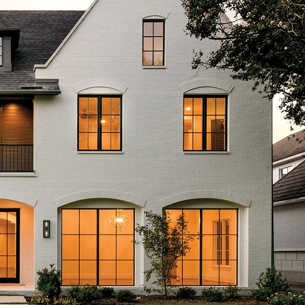 Exterior view of a modern two-story white brick house with black-framed, grid-style windows glowing with warm interior lighting at dusk.